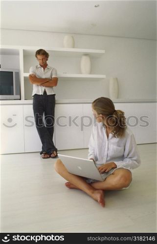Young woman sitting on the floor holding a laptop with a young man standing behind her