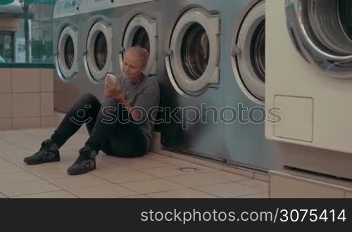 Young woman sitting on the floor in the laundry and passing the time with smart phone