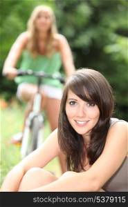 Young woman sitting on the grass with her friend on a bike in the background