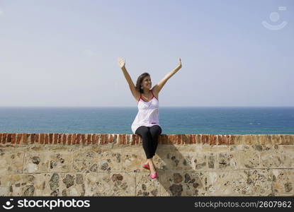 Young woman sitting on the ledge with her arms raised and smiling