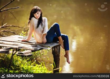 Young woman sitting on the old bridge barefoot. Summer, green forest, lake and air in the evening bring the mood to relax, meditate, enjoy the sounds of nature and dream. Young woman sitting on the old bridge barefoot. Summer, green forest, lake and air in the evening bring the mood to relax, meditate, enjoy the sounds of nature and dream.