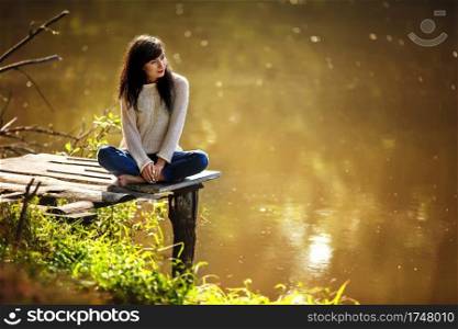 Young woman sitting on the old bridge barefoot. Summer, green forest, lake and air in the evening bring the mood to relax, meditate, enjoy the sounds of nature and dream. Young woman sitting on the old bridge barefoot. Summer, green forest, lake and air in the evening bring the mood to relax, meditate, enjoy the sounds of nature and dream.