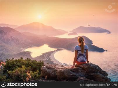 Young woman sitting on the top of rock and looking at the seasho. Young woman sitting on the top of rock and looking at the seashore and mountains at colorful sunset in summer. Landscape with girl, sea, mountain ridges and orange sky with sun. Oludeniz, Turkey.