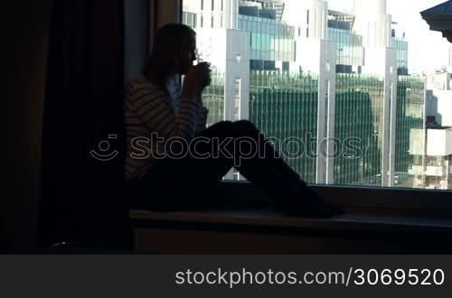 Young woman sitting on the windowsill with mug of tea or coffee and enjoying city panorama