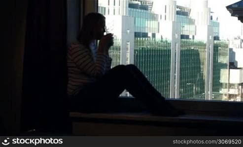 Young woman sitting on the windowsill with mug of tea or coffee and enjoying city panorama