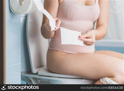 Young woman sitting on toilet and tearing off paper tissue