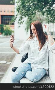 Young woman sitting on wall taking smartphone selfie