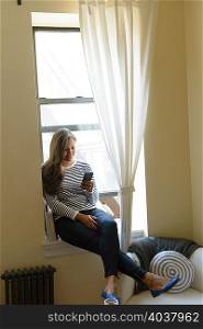 Young woman sitting on window sill using smartphone