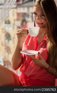 Young woman sitting on windowsill looking through window enjoying her free time, relaxing while drinking coffee or tea from cup.. Woman looking through window, relaxing drinking coffee