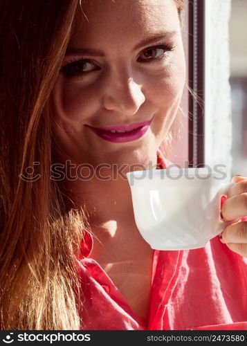 Young woman sitting on windowsill looking through window enjoying her free time, relaxing while drinking coffee or tea from cup.. Woman looking through window, relaxing drinking coffee