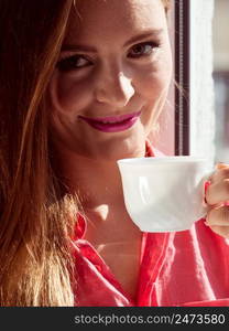 Young woman sitting on windowsill looking through window enjoying her free time, relaxing while drinking coffee or tea from cup.. Woman looking through window, relaxing drinking coffee