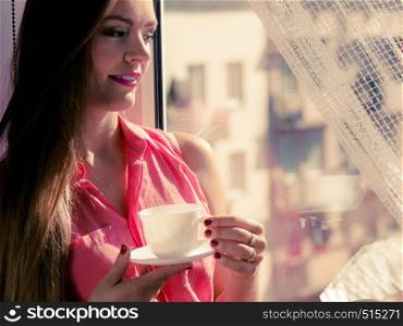Young woman sitting on windowsill looking through window enjoying her free time, relaxing while drinking coffee or tea from cup.. Woman looking through window, relaxing drinking coffee