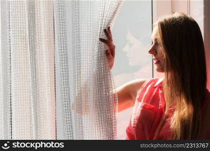 Young woman sitting on windowsill looking through window enjoying her free time, relaxing.. Woman looking through window, relaxing