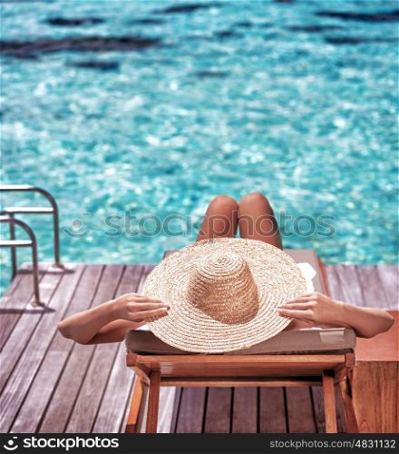 Young woman sitting on wooden pier on the sea wearing hat and taking sun bath, enjoying perfect summer day, traveling and luxury vacation concept
