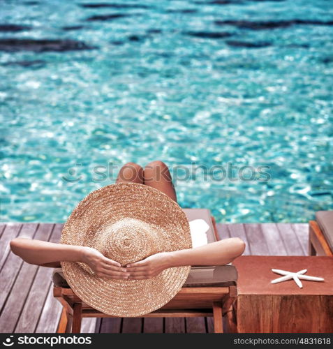 Young woman sitting on wooden pier on the sea wearing hat and taking sun bath, enjoying perfect summer day, traveling and luxury vacation concept