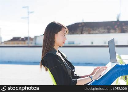 Young woman sitting with feet up on city roof terrace typing on laptop