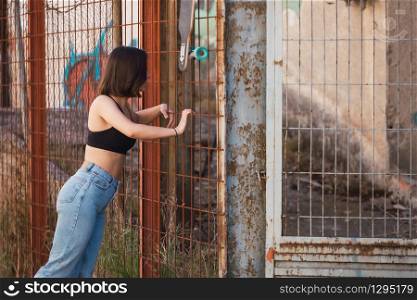 Young woman skater look inside through a fence in a ancient industrial street