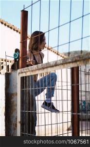 Young woman skater trying to climb over the fence look inside in a ancient industrial street