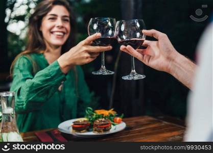 Young woman smiling and enjoying romantic vegetarian dinner served in restaurant. Toasting with red wine on a date. Sitting at wooden table, wearing colorful green outfit.  