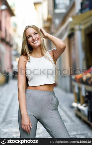 Young woman smiling in urban background. Blond girl with straight hairstyle wearing casual clothes in the street.