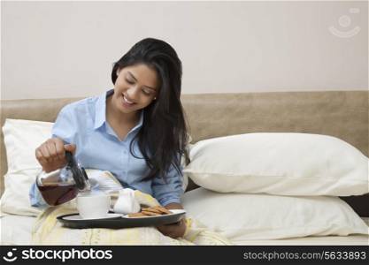 Young woman smiling while pouring tea in cup while sitting in bed