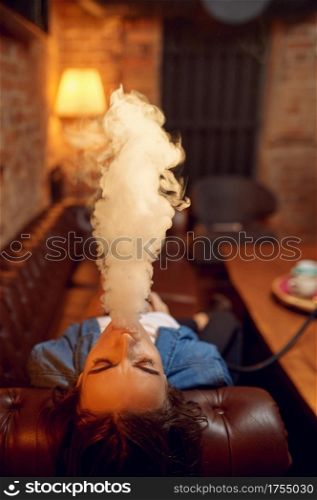 Young woman smokes on sofa in hookah bar, mesmerizing cloud. Shisha smoking, traditional bong culture, tobacco aroma for relaxation, rest with hooka. Young woman smokes on sofa in hookah bar