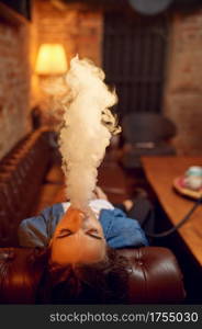 Young woman smokes on sofa in hookah bar, mesmerizing cloud. Shisha smoking, traditional bong culture, tobacco aroma for relaxation, rest with hooka. Young woman smokes on sofa in hookah bar