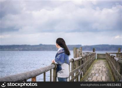 Young woman standing alone, on an old wooden bridge, thinking and gazing the horizon in the afternoon, over the Chiemsee lake, in Bavaria, Germany.