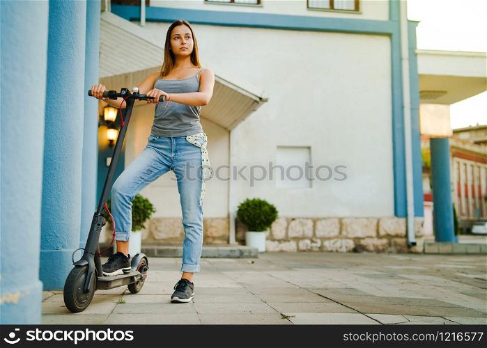 Young woman standing by electric kick scooter by the building in the town city waiting in summer day alternative transport