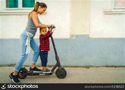 Young woman standing by the electric kick scooter board on the pavement in front of the wall of some house in the town riding driving her little small son or brother boy