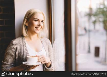Young woman standing by the window with a cup of coffee, looking outside and smiling. Natural outdoor light.. Woman with a cup of coffee standing by the window.