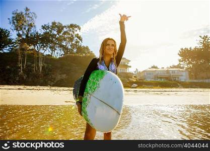 Young woman standing in sea, looking out to sea, holding surfboard, gesturing with hand