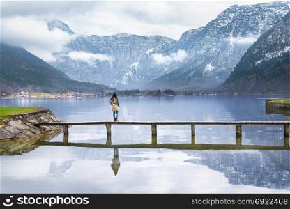 Young woman standing on a wooden bridge over the Hallstatter lake, enjoying the silence and the view of the Austrian Alps mountains reflected in the water.