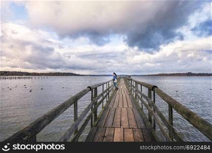 Young woman standing on an old wooden bridge, meditating and admiring the view of the Chiemsee lake, in the afternoon, located near Rosenheim, Germany.