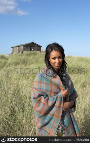 Young Woman Standing On Beach Wrapped In Blanket With Beach Hut In Distance