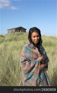 Young Woman Standing On Beach Wrapped In Blanket With Beach Hut In Distance