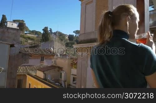Young woman standing on outdoor balcony. She drinking hot tea and enjoying city view
