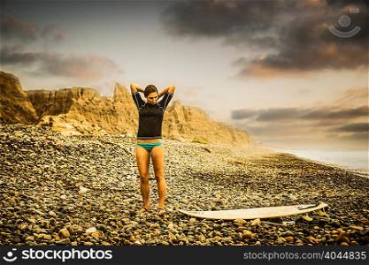 Young woman standing on pebble beach looking down at surfboard
