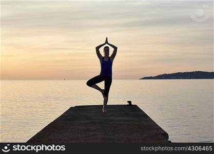 Young woman standing on pier, in yoga position, at sunset