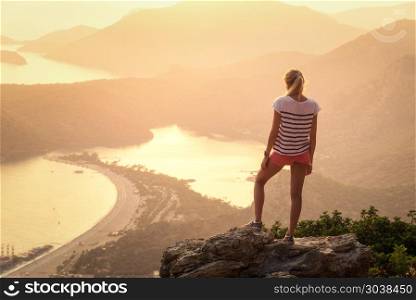 Young woman standing on the top of rock and looking at the seashore and mountains at colorful sunset in summer. Landscape with girl, sea, mountain ridges and orange sunlight. Travel. Oludeniz, Turkey. Landscape with woman, sea, mountain ridges and orange sky