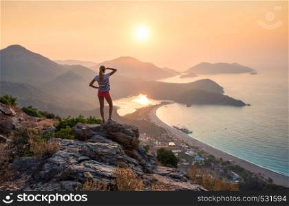 Young woman standing on the top of rock and looking at the seashore and mountains at colorful sunset in summer. Landscape with girl, sea, mountain ridges and orange sky with sun. Oludeniz, Turkey. Landscape with girl, sea, mountain ridges and orange sky