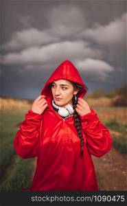 Young woman standing with raincoat and headphones on the field in a rainy day
