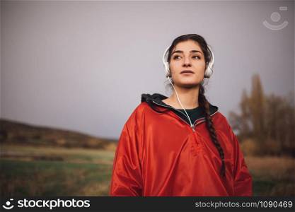 Young woman standing with raincoat listening to music with headphones on field in a rainy day