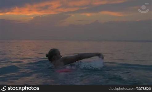 Young woman swims away in the sea at sunset. Magical view of evening cloudy sky and calm sea