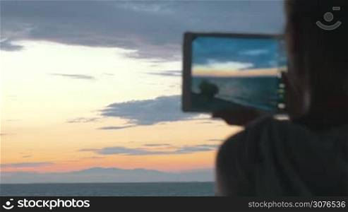 Young woman takes photos of the beautiful scenery of the sea and evening sky with her tablet at sunset