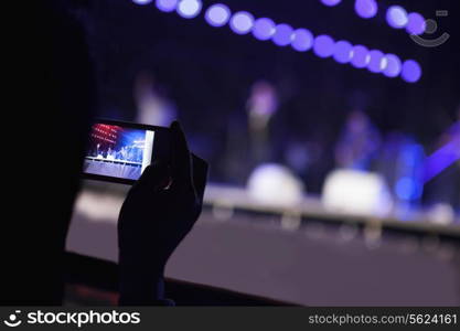 Young woman taking a photography with her smart phone at an indoor concert, over the shoulder view