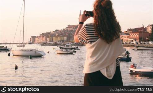 Young woman taking a picture of Rovinj marina and city view with boats and St. Euphemia cathedral at sunset. Istria, Croatia.. Young woman taking a picture of Rovinj