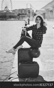 Young woman taking photographs with a vintage camera sitting in a harbour. Girl wearing plaid shirt, blue jeans and sun hat. Black and white photograph.