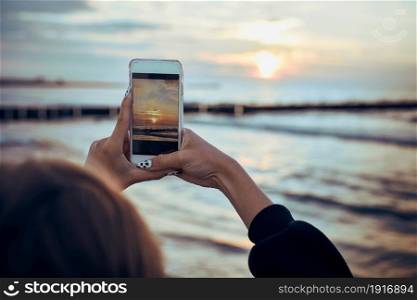 Young woman taking photos of sunset over sea using smartphone during summer trip. Rear view of girl holding smartphone and taking picture