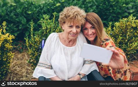 Young woman taking selfie with her older mother in a wheelchair. Girl taking selfie with her mother in wheelchair